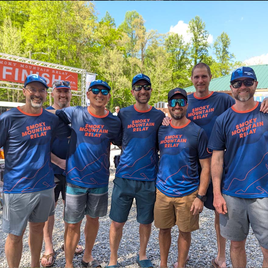Group of men in 'Smoky Mountain Relay' shirts posing outdoors with trees and a finish line sign in the background.