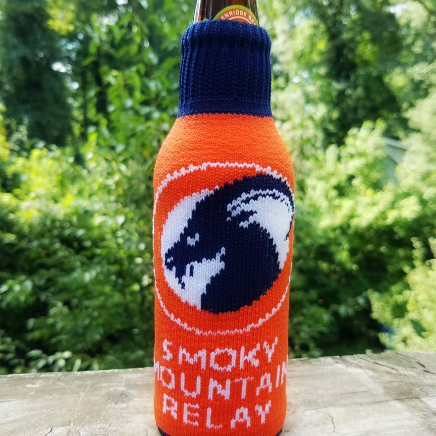 Bottle with orange and blue knitted cover featuring SMR ram logo and text, set against a blurred green outdoor background.