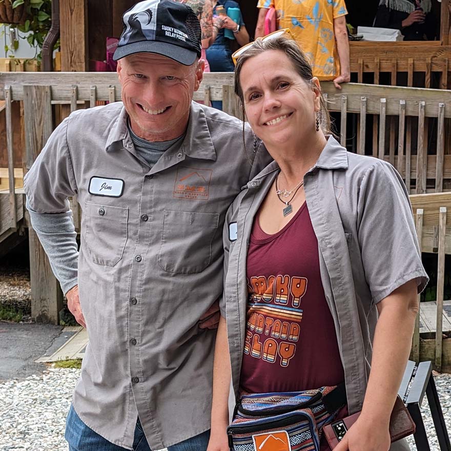 Two people standing together outdoors, smiling at the camera, one wearing a maroon tank top with colorful 'Smoky Mountain Relay' text design.