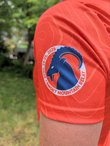 Orange shirt with Smoky Mountain Relay logo on the left sleeve on a blurred outdoor background
