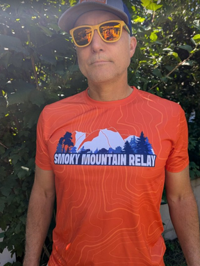 Person wearing an orange 'Smoky Mountain Relay' t-shirt with sunglasses and a cap outdoors.