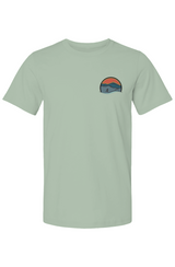 front of light green t-shirt and on left chest a small Run Asheville logo featuring a circular emblem with layered mountain ridges in teal and slate blue tones beneath a warm red-orange sky with a golden horizon. A silhouette of a runner strides across the foreground, with a flowing light teal wave element at the base of the circle and the URL "RUNASHEVILLE.COM" arced across the lower interior.

