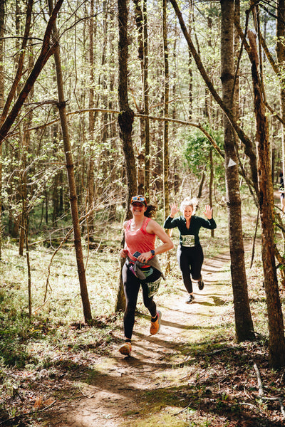 Two runners on a forest trail, one in a pink shirt and black pants, the other in a black jacket and black pants.