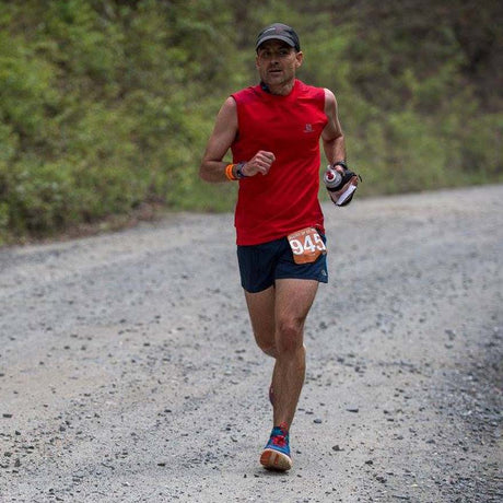Man running on a gravel road wearing a red sleeveless shirt and black shorts with a race number.